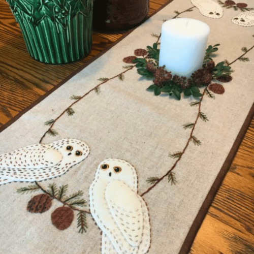 Carried Away Designs Linen-colored table runner with 2 snowy owls at each end and evergreen on wooden table with white candle in center
