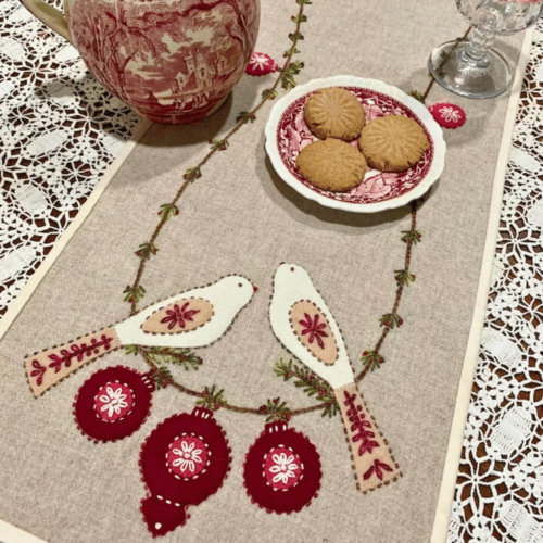 Carried Away Designs linen-colored table runner with two birds and red ornaments at each end with vintage red pitcher and plate of cookies