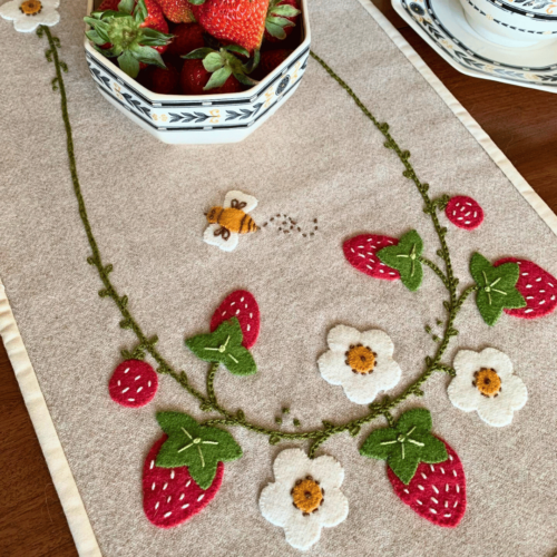 Carried Away Designs linen-colored table runner with strawberries and white flowers on wooden table with bowl of strawberries in center