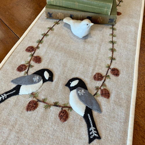 Carried Away Designs linen-colored table runner with two chickadees at each end with vintage red pitcher and stack of books in center