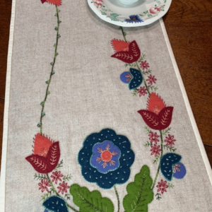Carried Away Designs linen-colored table runner with Jacobean red and blue floral pattern at each end with a cup and saucer in the center