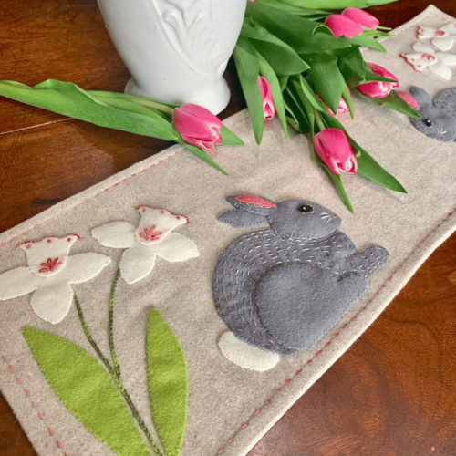 Carried Away Designs linen-colored table runner with gray rabbit and white daffodils at each end with white vase and pink tulips