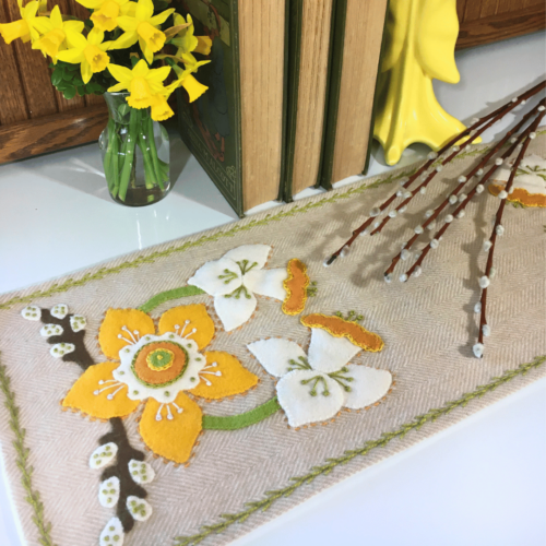 Carried Away Designs linen-colored table runner with white and yellow daffodils at each end with books and yellow flowers in the background