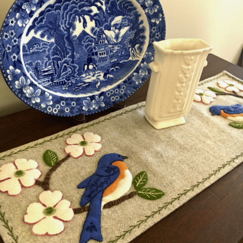 Carried Away Designs linen-colored table runner with a bluebird at each end with white flowers on wooden table with blue plate in background